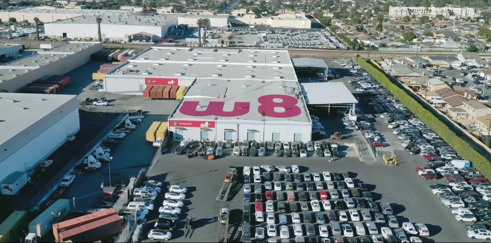 В центре фото склад с большим логотипом W8 на крыше и логотипами поменьше над несколькими входами. Вокруг склада парковка, на которой припарковано много автомобилей.